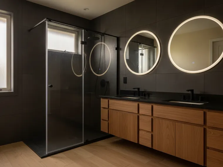 Moody master bathroom idea: charcoal large-format tile, floating walnut double vanity with a black top, two backlit round mirrors, black-framed glass shower and a pale wood floor