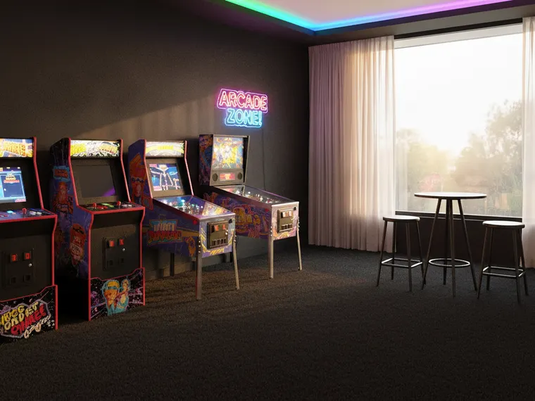 Game room idea: three upright arcade cabinets and a pinball machine along a black wall under a neon sign, with a small bar table and colour strips at the ceiling