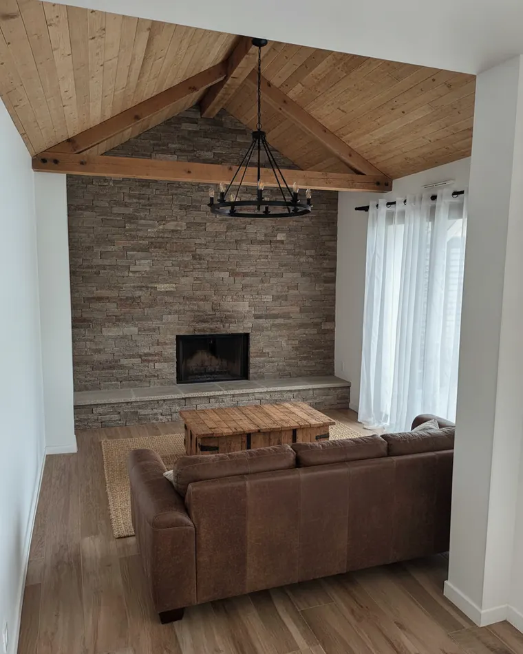 Barndominium living room idea: floor-to-ceiling stacked stone fireplace, wood plank vaulted ceiling with beams, leather sectional and iron chandelier