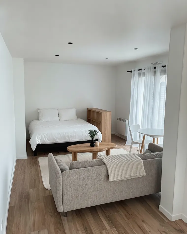 Small studio apartment layout idea: a double bed with white bedding against the far wall, a low oak bookshelf between the bed and a grey sofa, a round coffee table and a small round dining table with two chairs by the window