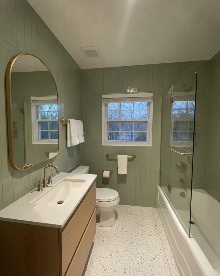 Master bathroom design idea: sage green vertical tile on the walls, white oak vanity with an arched brass mirror, brass fixtures, glass screen on a white tub and a terrazzo floor