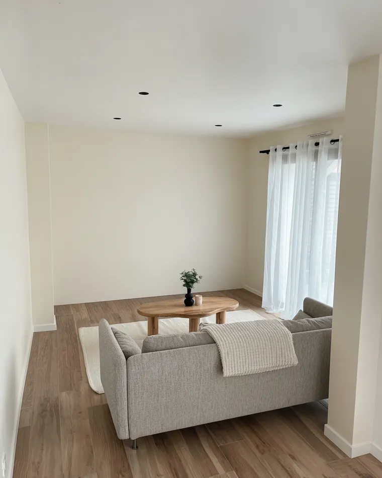 Neutral living room paint colour: warm cream walls in a bright room with a grey sofa and sheer curtains