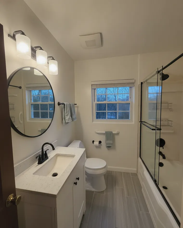 Cheapest master bathroom idea in a three-piece: walls painted soft white, a white vanity with a stone top, a round black-framed mirror, matte black fixtures, a black-framed glass panel on the tub and grey wood-look floor tile