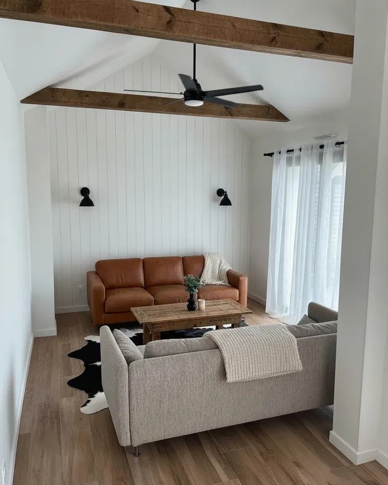 Barndominium living room idea: vertical white shiplap wall, a dark ceiling beam with a black fan, cognac leather sofa, cowhide rug and black sconces