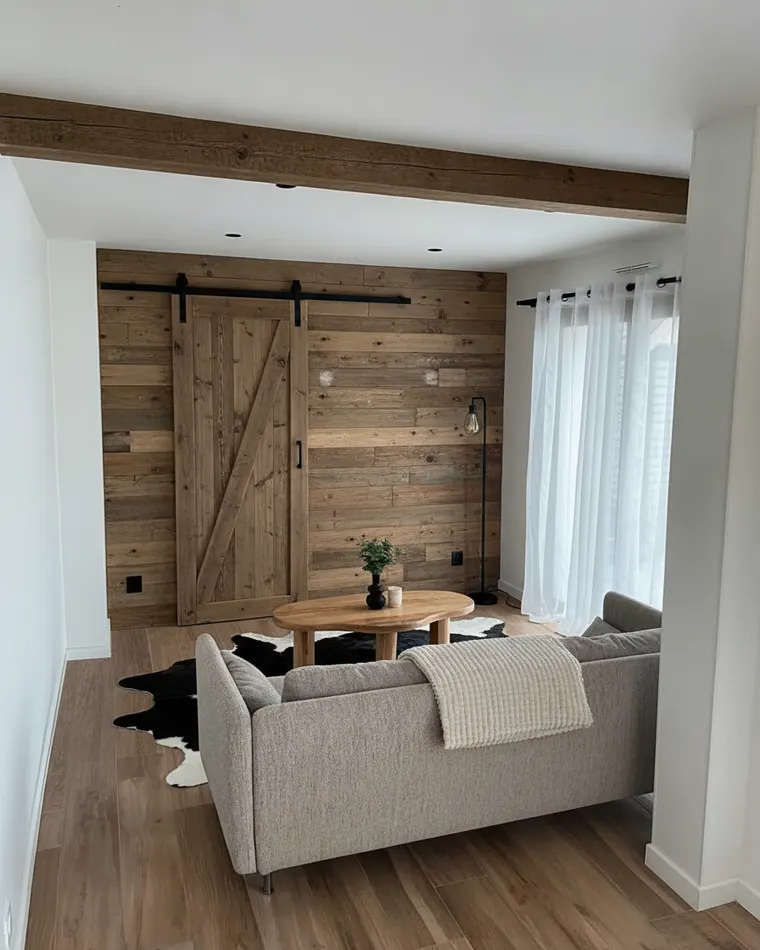 Barndominium living room idea that keeps the layout: reclaimed wood plank accent wall, sliding barn door, black floor lamp, cowhide rug and a ceiling beam
