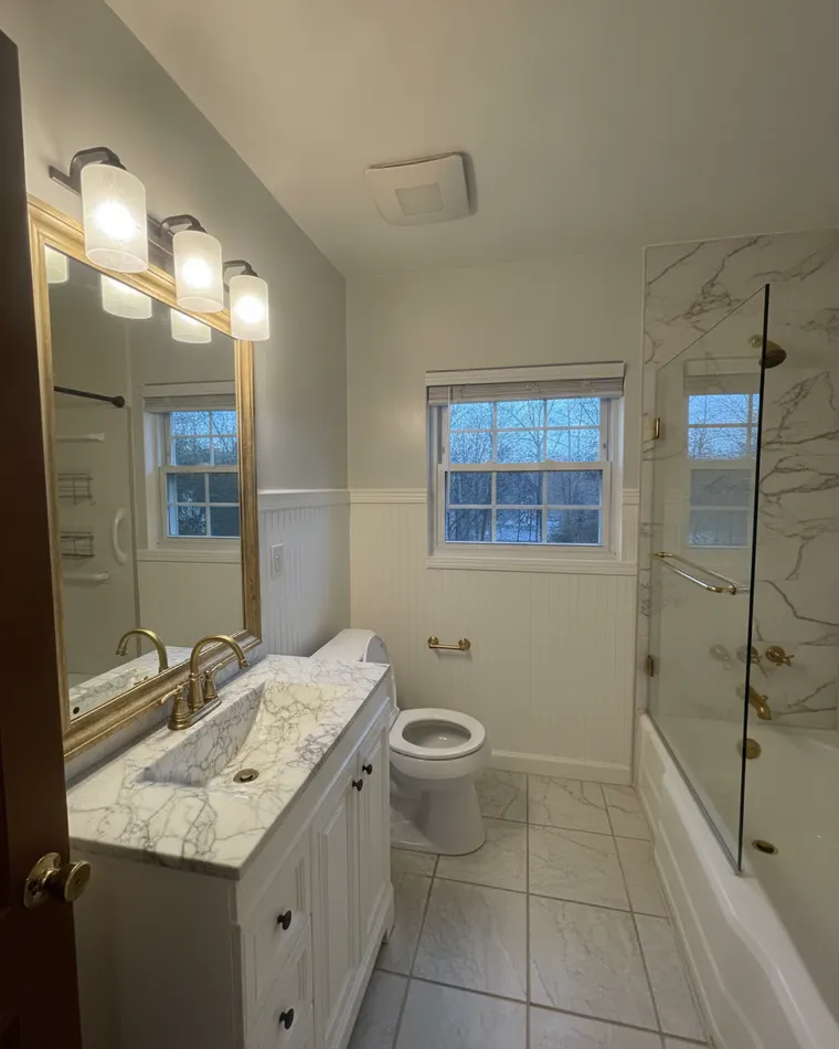 Classic master bathroom idea: white marble tile around the tub, white beadboard wainscoting, white vanity with a marble top, gold-framed mirror, brass fixtures and a glass screen