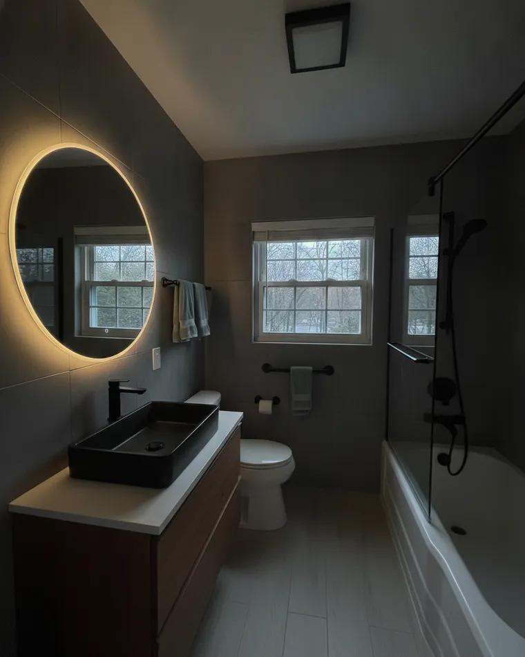 Dark master bathroom idea in a narrow three-piece: matte charcoal wall tile, walnut floating vanity with a black stone basin, backlit round mirror, black-framed glass screen and pale wood floor