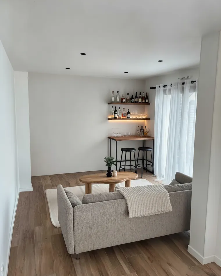 Small home bar idea: two lit wooden shelves of bottles and glassware in a living room corner above a slim black bar table with two stools
