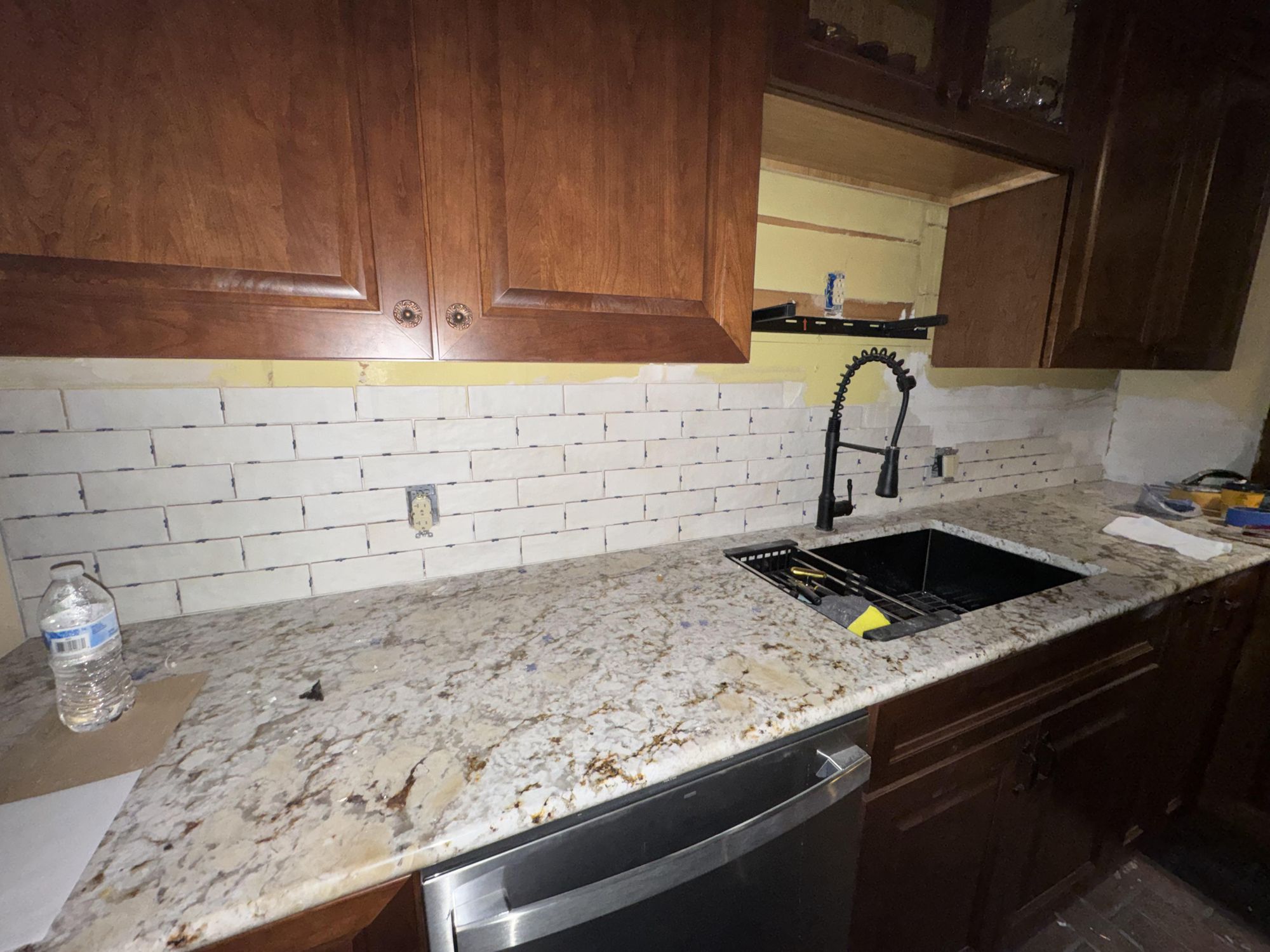 White subway tile backsplash mid-install over cherry cabinets and busy granite, with messy unfinished grout that makes the wall look like a mistake